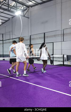 Divers joueurs de padel marchant à travers le filet sur le court violet dans la salle de sport avec escrime, raquettes Banque D'Images