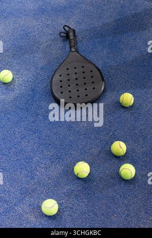 Raquette de padel noir reposant sur la surface du court texturée bleue parmi les balles de tennis jaunes Banque D'Images