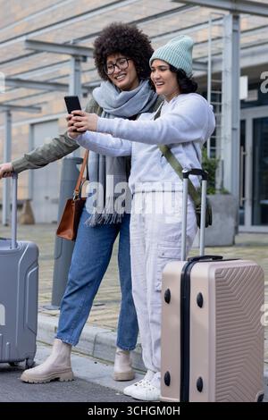 Diverses amies féminines debout à l'extérieur de l'entrée de l'aéroport vérifiant smartphone, valises roulantes Banque D'Images