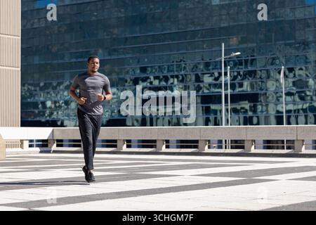Homme afro-américain jogging dans l'équipement sur la plaza par bâtiment de verre, espace copie Banque D'Images