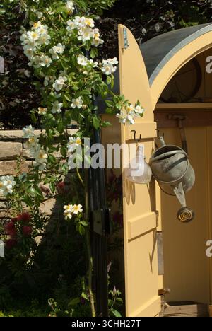 Un cabanon de jardin confortable peint dans des stands jaunes parmi des fleurs vibrantes avec un arrosoir suspendu à proximité sous la lumière douce de l'après-midi. Banque D'Images