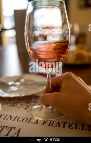 Gros plan d'un verre de vin rosé tenu dans une main, avec un arrière-plan flou mettant en valeur un cadre de restaurant et une vue partielle d'un panneau de lecture Banque D'Images