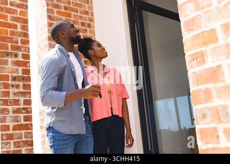 Couple heureux tenant les clés, debout à l'extérieur de la nouvelle maison, l'air excité et plein d'espoir Banque D'Images