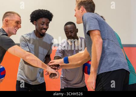 Divers coéquipiers masculins empilant les mains dans une salle de sport tenant des balles de volley, portant des écouteurs Banque D'Images