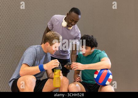 Divers partenaires masculins vérifiant smartphone sur banc à la salle de gym avec bouteille d'eau écouteurs de volley-ball Banque D'Images