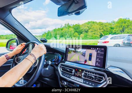Conducteur tenant le volant à backgroundю intérieur de la BMW iX3 M électrique avec affichage montrant la caméra de stationnement et le paysage en Suède. Banque D'Images