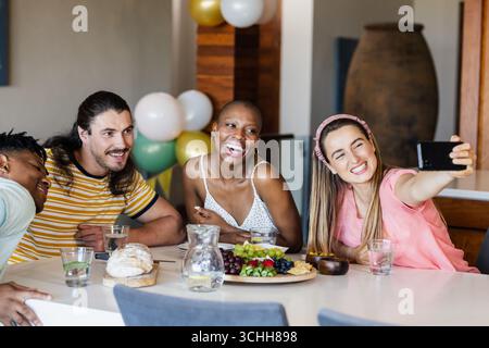 Des amis divers appréciant le déjeuner ensemble, souriant et prenant selfie à la table à manger, à la maison Banque D'Images