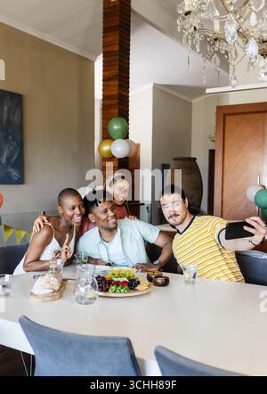 Amis divers prenant selfie à la maison, souriant autour de la table avec des collations et des boissons Banque D'Images