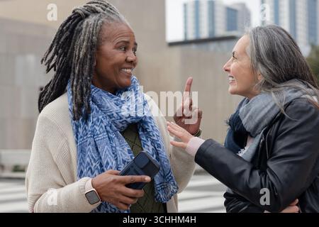 Diverses amies féminines discutant sur le passage de la ville portant des foulards à l'aide d'un smartphone Banque D'Images