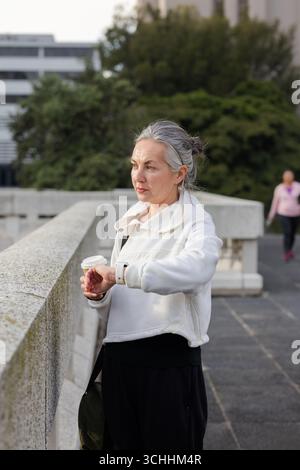 Femme senior avec tasse à café à emporter tout en vérifiant smartwatch sur la passerelle urbaine sur le toit Banque D'Images