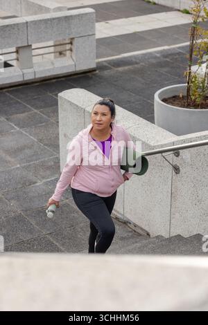 Femme grimpant l'escalier par la main courante dans la place urbaine portant tapis de yoga, bouteille d'eau, espace de copie Banque D'Images