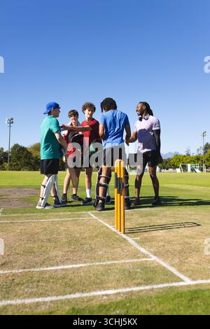 Divers coéquipiers masculins debout sur un terrain de cricket à côté de souches tenant des chauves-souris, des casques et des coussinets pour les jambes Banque D'Images