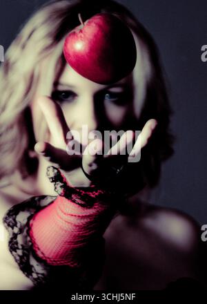 Une femme aux cheveux blonds et aux gants en résille rouge tend la main à une pomme rouge flottant devant son visage. Banque D'Images
