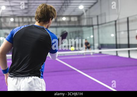 Deux hommes jouant au paddle-tennis sur un court violet avec des raquettes et balle jaune sur le filet, espace copie Banque D'Images