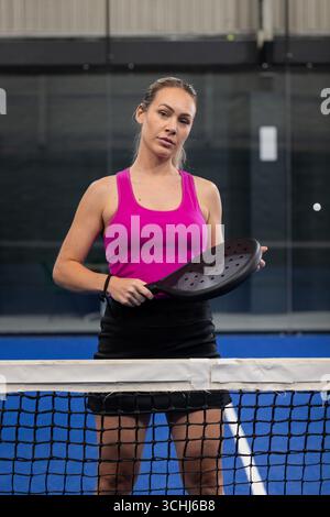 Joueuse debout derrière un filet de padel avec une raquette noire sur le court portant un haut rose et une jupe noire Banque D'Images