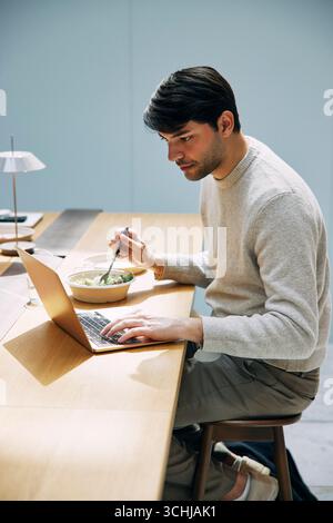 Jeune freelance utilisant un ordinateur portable tout en mangeant le déjeuner assis sur le tabouret à la table dans la cafétéria de bureau Banque D'Images