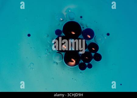 Grappe de gouttelettes d'huile foncées suspendues dans un liquide bleu, entourées de sphères plus petites sur un fond cyan, photographiées en macro avec un niveau de détail élevé. Banque D'Images