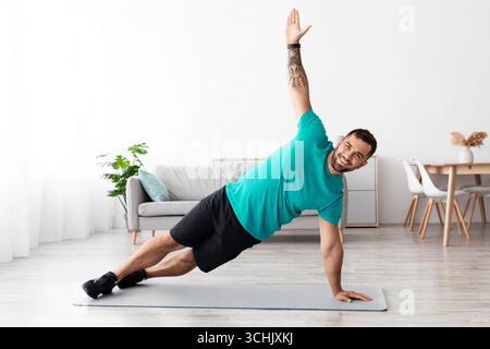 Souriant joyeux athlète européen millénaire faisant des planches de côté sur le tapis dans le salon minimaliste Banque D'Images