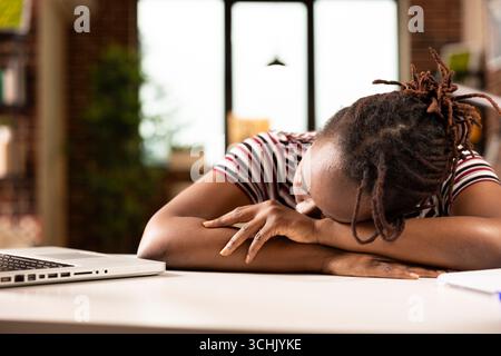 Femme entrepreneure afro-américaine dormant au bureau après avoir surtravaillé sur un ordinateur portable. Femme d'affaires noire fatiguée reposant la tête sur ses bras, faisant la sieste pour récupérer de longues heures à la maison. Banque D'Images