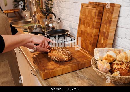 Femme tranchant du pain multigrain sur une planche à découper en bois dans une cuisine domestique confortable Banque D'Images