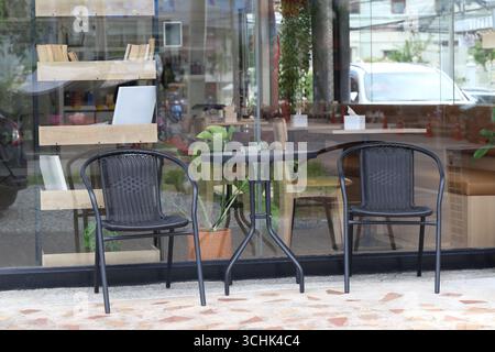 Un charmant coin salon extérieur avec deux chaises noires et une table ronde à côté d'une vitrine, ornée de plantes, créant un accueil et rela Banque D'Images
