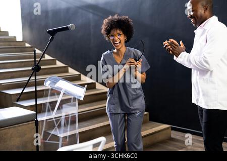Recevoir des applaudissements sur scène, femme en gommage souriant, tenant des lunettes, à la salle de conférence Banque D'Images