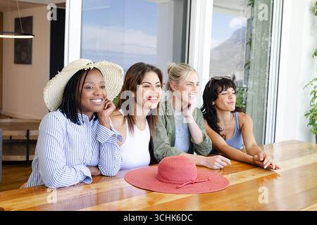 Groupe diversifié d'amies féminines diverses assis à table, souriant et profitant d'une journée ensoleillée, à la maison Banque D'Images