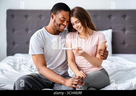 Portrait d'un heureux couple multiracial tenant un test positif de grossesse, assis sur le lit, se tenant et appréciant les bonnes nouvelles Banque D'Images