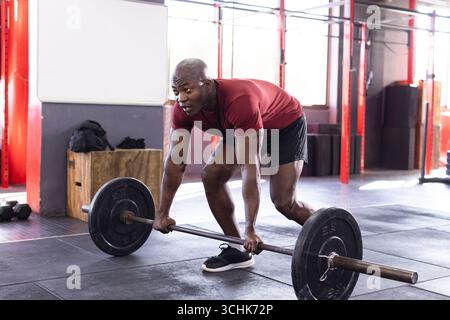 Homme afro-américain portant un tee-shirt rouge short noir pliant, saisissant barre à la salle de gym avec poutres Banque D'Images