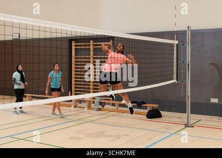 Diverses coéquipières féminines pratiquant le volley-ball dans la salle de gym, sautant par-dessus le filet par le banc, sac de sport Banque D'Images