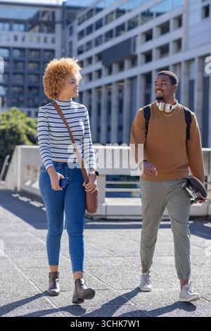 Divers amis marchant à côté de la rampe en béton sur la passerelle urbaine portant skateboard, smartphone Banque D'Images