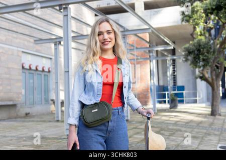 Femme dans la cour portant une veste en denim tenant un chapeau de soleil de paille portant un sac de bandoulière vert Banque D'Images