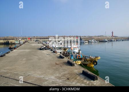 De petits bateaux de pêche s'attachent le long des deux côtés d'une jetée en béton au port de Sagye, avec des filets et des bouées empilés sur les ponts et un phare rouge à travers le bassin. Banque D'Images
