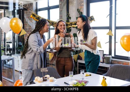Collègues féminins divers célébrant leur anniversaire au bureau, appréciant le gâteau et riant ensemble Banque D'Images