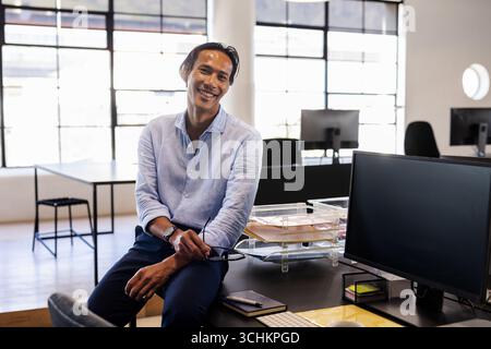 Homme d'affaires souriant dans le bureau moderne appuyé sur le bureau, tenant des verres, espace de copie Banque D'Images