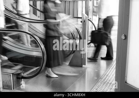 Femme d'affaires floue et homme avec escalator de sortie de mallette dans une scène monochrome capturant le mouvement, la vie moderne de la ville, et des moments fugaces Banque D'Images