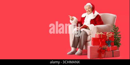 Femme senior habillée comme le Père Noël assis dans un fauteuil et pointant vers quelque chose tout en lisant le livre sur fond rouge avec un espace pour le texte Banque D'Images