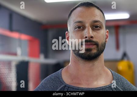 Homme indien debout dans la salle de gym faisant face à la caméra avec sac lourd, barrière en maille, panneaux muraux rembourrés Banque D'Images