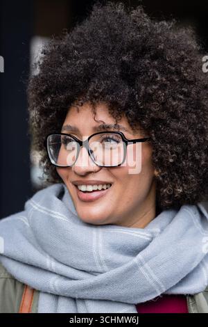 Femme portant des lunettes à cadre noir, anneau de nez, foulard gris souriant, regardant à gauche près de l'entrée Banque D'Images
