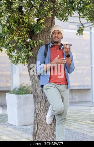 Homme afro-américain appuyé contre l'arbre dans la plaza tenant smartphone, portant des écouteurs Banque D'Images