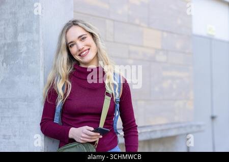 femme des années 20 s'appuyant sur la colonne au trottoir tenant le téléphone dans une combinaison en denim, sac vert, espace de copie Banque D'Images