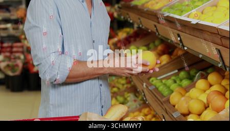 magasinier en chemise rayée choisissant orange dans l'allée des fruits et légumes frais avec étagères, chariot rouge contenant des baguettes Banque D'Images
