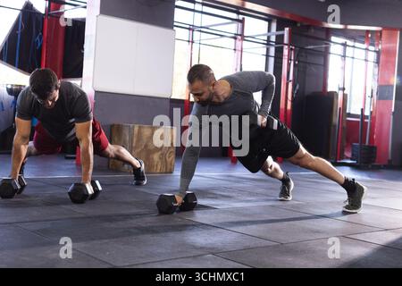 Divers partenaires d'entraînement masculins effectuant des rangées de planches sur tapis dans le gymnase à l'aide d'haltères hexagonaux Banque D'Images