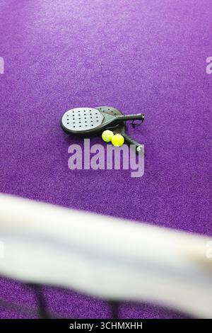 Deux raquettes de padel noir reposent sur un terrain en gazon violet à côté de boules jaunes à côté du filet flou Banque D'Images