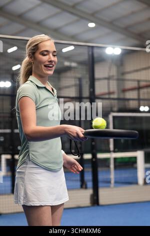 Athlète féminine tenant une raquette de padel équilibrant une balle de tennis sur un court de padel près d'une clôture métallique, filet Banque D'Images