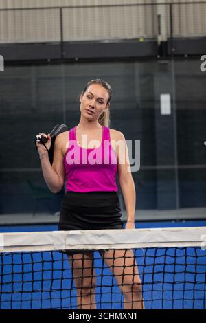 Joueuse debout derrière un filet de padel sur un court bleu avec des murs de verre tenant la raquette sur l'épaule Banque D'Images