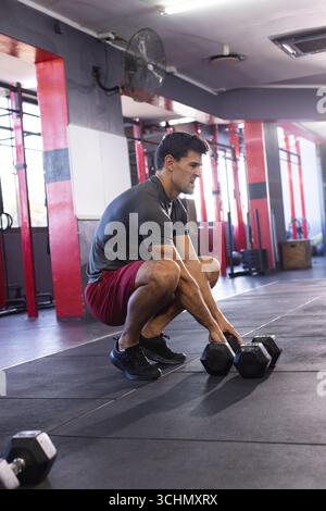 Homme accroupissant à la salle de gym saisissant des haltères hexagonaux sur des tapis en caoutchouc près des casiers de squat, des boîtes plyométriques Banque D'Images