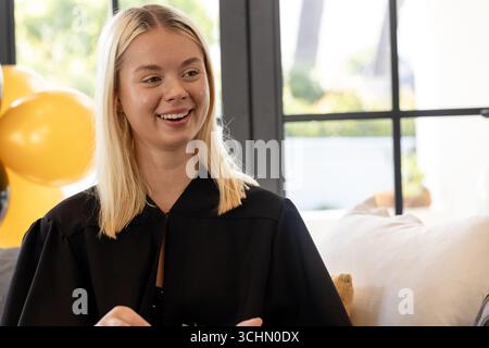 Jeune femme en robe de graduation souriant à la maison pendant la célébration Banque D'Images