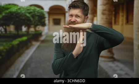 Homme avec une longue barbe rouge fait un geste de temps hors du temps avec les mains sur la rue pavée à côté des arches historiques ; pause. Banque D'Images