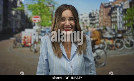 Femme souriante en chemise bleue avec des lunettes et un large sourire du doigt pointant vers le canal ensoleillé sur la rue ; joie insouciante. Banque D'Images